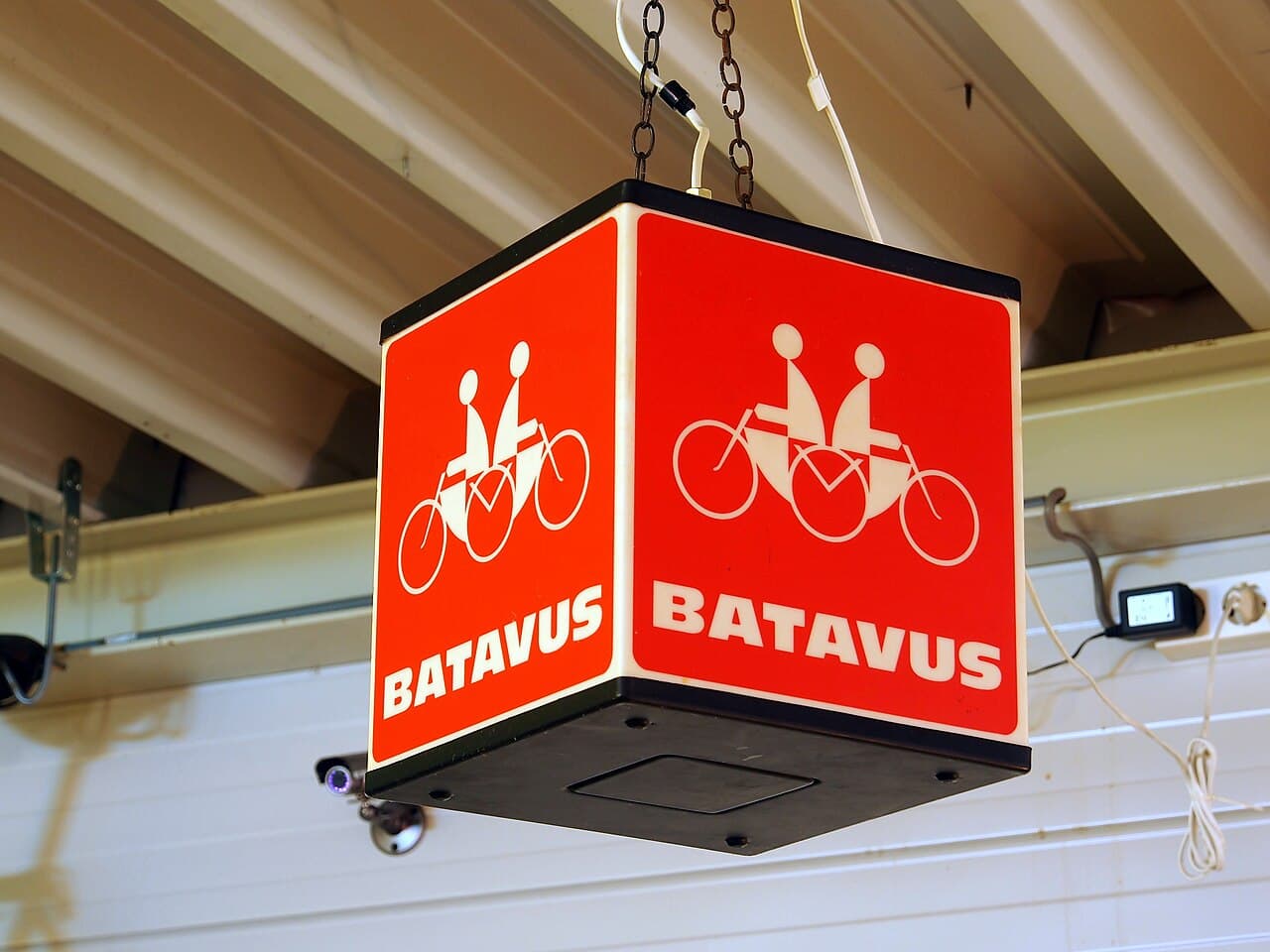 Rode, kubusvormige Batavus-lichtreclame met het logo van twee fietsers op een tandem, aan kettingen opgehangen onder een plafond