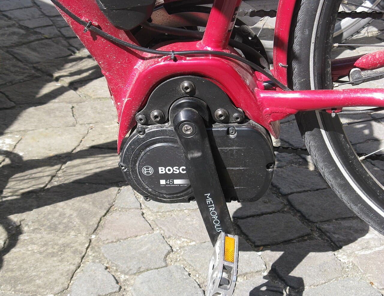 Zwarte Bosch-middenmotor onderin het rode frame van een e-bike, met crank en pedaal, gefotografeerd op straatstenen