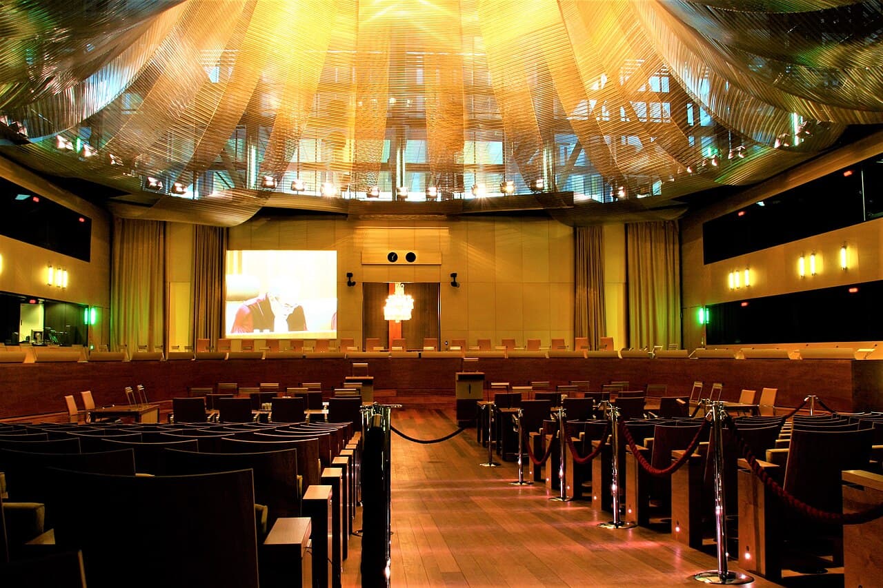 Grote rechtszaal van het Hof van Justitie van de Europese Unie in Luxemburg, met lege houten stoelen, een scherm boven de rechterstafel en een gouden plafond