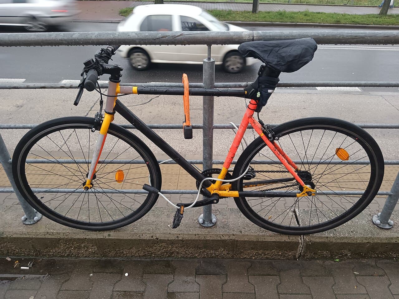 Fiets met een oranje U-slot aan de stang en een kabelslot door het wiel, vastgezet aan een hek langs een straat