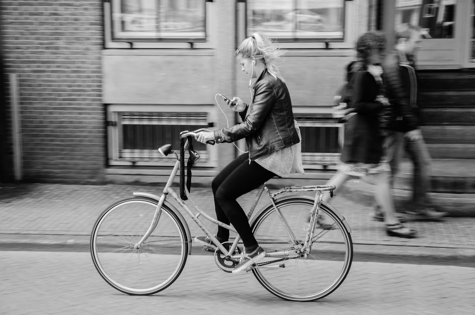 Zwart-witfoto van een fietsster in Amsterdam die al rijdend op haar telefoon kijkt, met oortjes in en voetgangers op de achtergrond