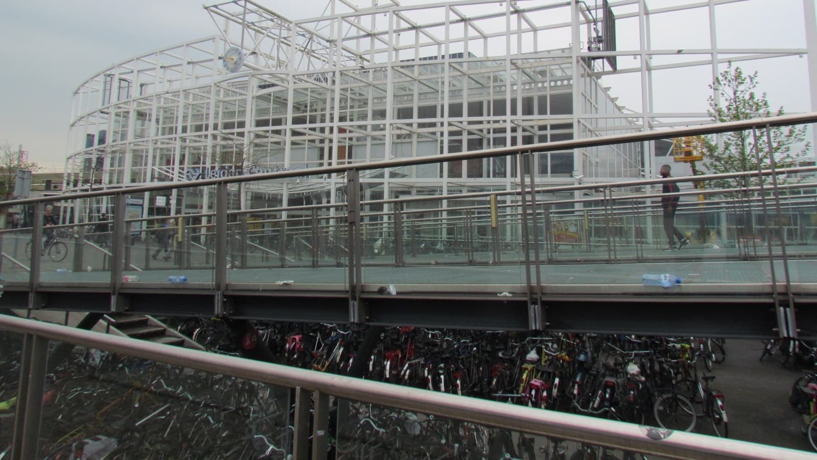 Volle fietsenstalling met honderden geparkeerde fietsen onder de loopbrug bij station Leiden Centraal, met het witte stalen frame van het stationsgebouw erachter