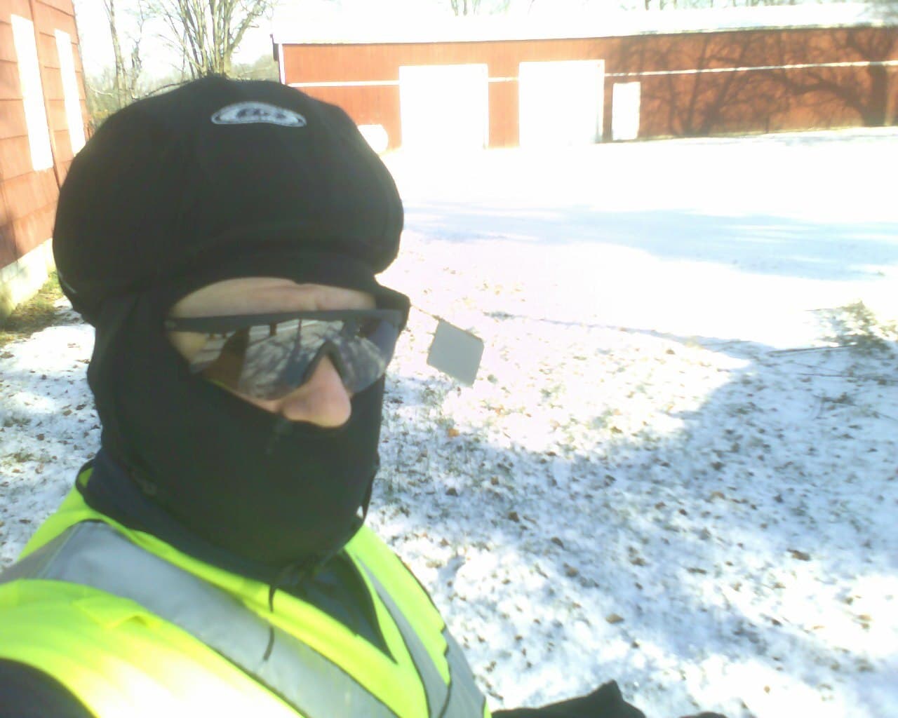 Fietser met zwarte bivakmuts, zonnebril en geel reflecterend hesje voor een besneeuwd erf in de winter