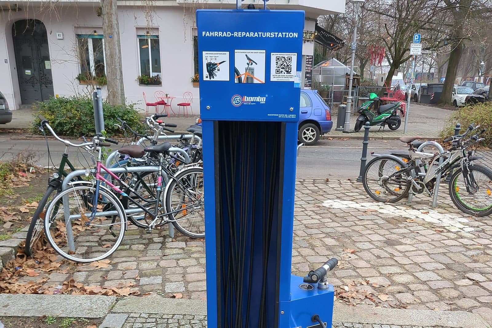 Blauwe openbare fietsreparatiezuil met pomp en gereedschap aan kabels op een kasseienplein, met geparkeerde stadsfietsen en een kinderfiets erachter
