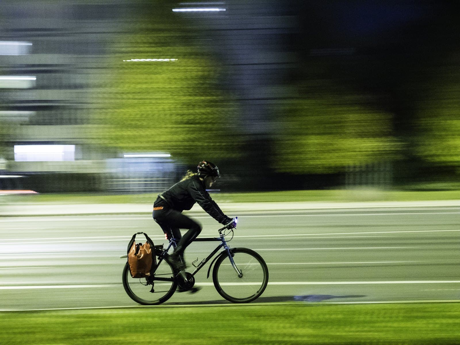 Fietser rijdt in het donker over een verlichte straat, met een wit lampje op het stuur en een rood lichtje achterop het frame; de achtergrond is bewogen doordat de camera meedraaide