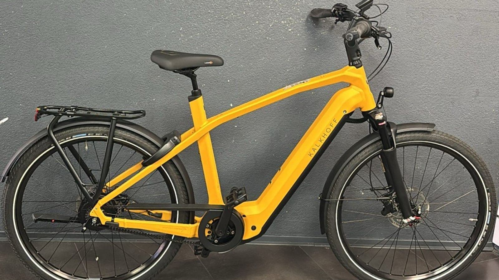 Kalkhoff Image 7.B Excite+ in matgeel, zijaanzicht in de winkel met Gates-riemaandrijving, Bosch-middenmotor, luchtgeveerde voorvork en bagagedrager