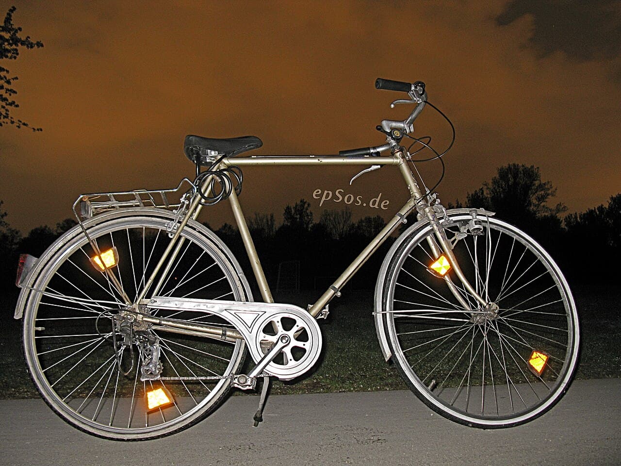 Oude herenfiets in het donker van opzij gefotografeerd, met oplichtende oranje reflectoren in voor- en achterwiel en een rood achterlicht op het spatbord