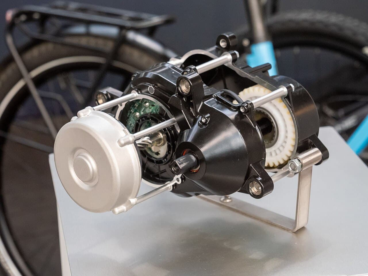 Opengewerkt model van een Panasonic GX-middenmotor op een beursstand, met zichtbare tandwielen en printplaat, voor een wazige e-bike (niet de GXM zelf)