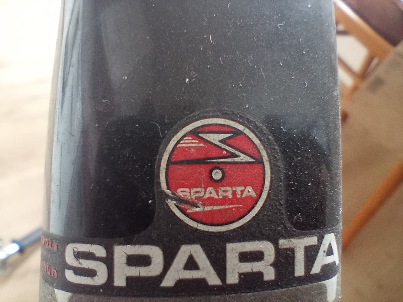 Rood rond Sparta-embleem met een wit S-logo boven de witte letters SPARTA op het zwarte frame van een oudere Sparta-fiets
