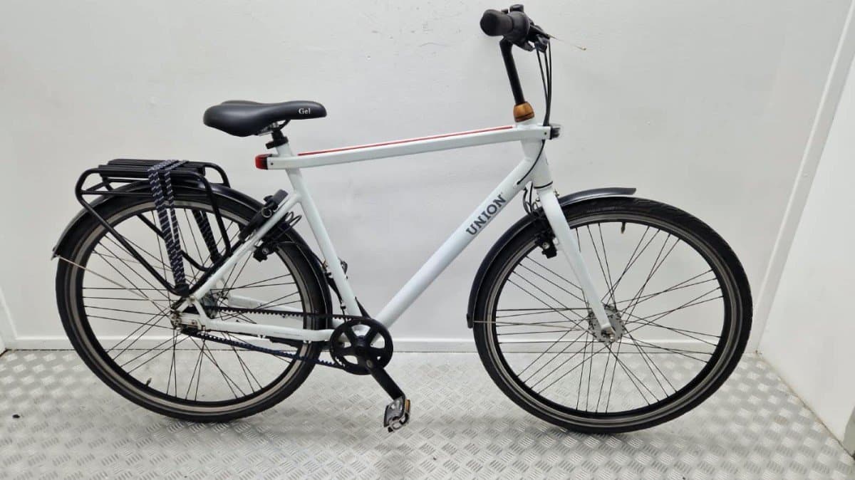 Witte Union Lite stadsfiets met zwarte riemaandrijving, spatborden, bagagedrager en 28 inch wielen, zijaanzicht tegen een witte wand