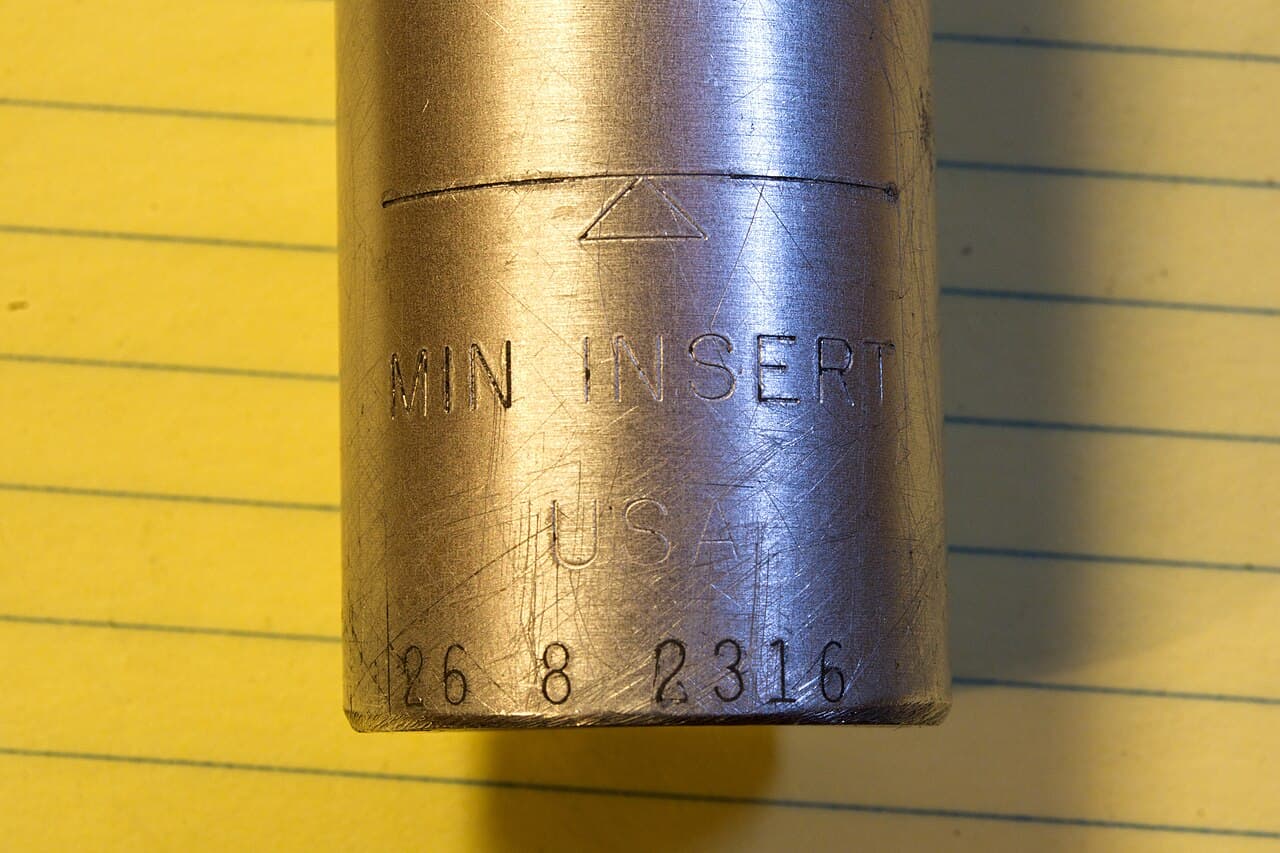 Close-up van een aluminium zadelpen met de gegraveerde markering MIN INSERT op gelinieerd geel papier