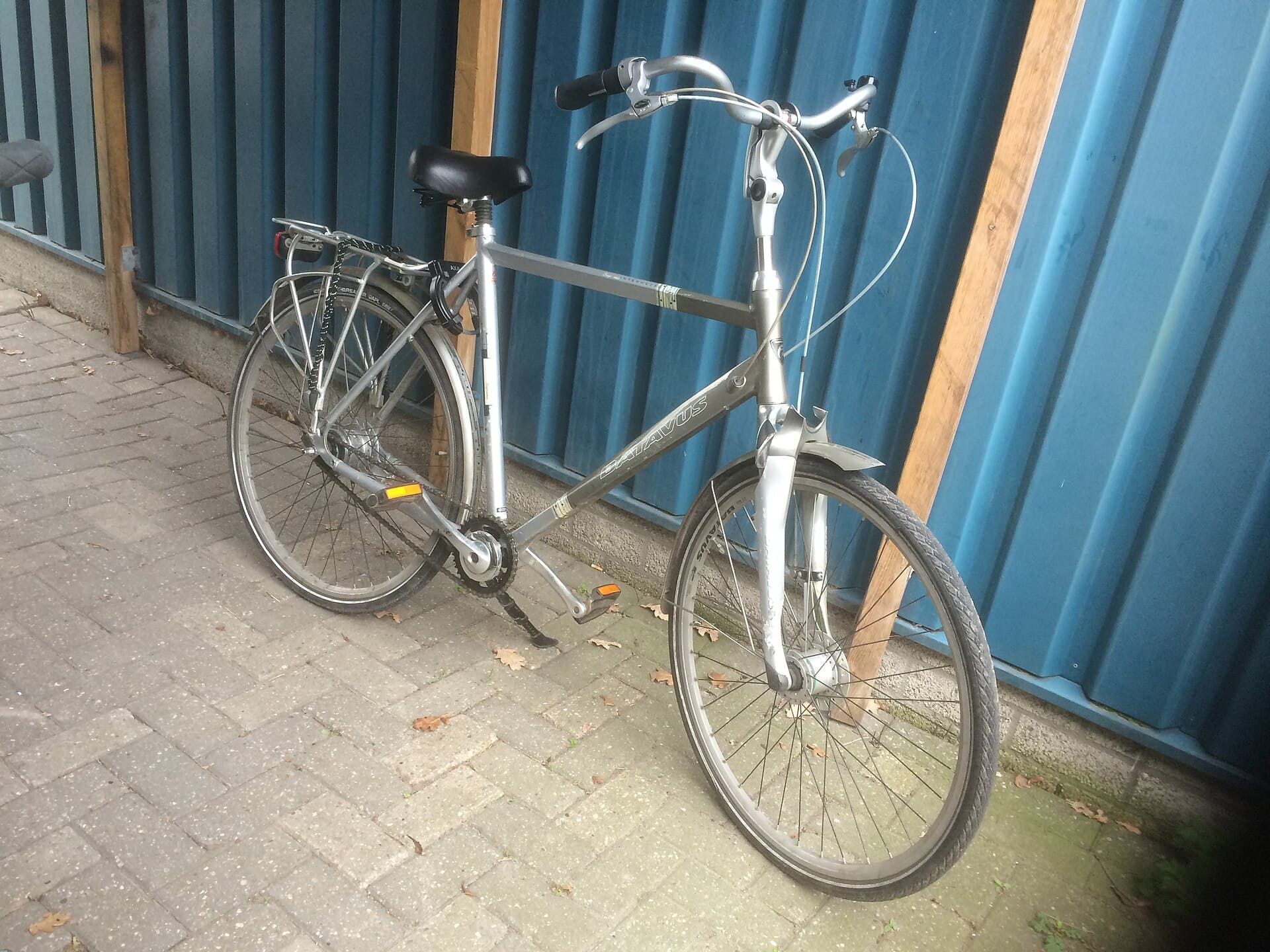 Zilvergrijze Batavus herenfiets met recht stuur, spatborden en bagagedrager, geparkeerd tegen een houten paal voor een blauwe golfplatenwand; de merknaam Batavus staat op de onderbuis