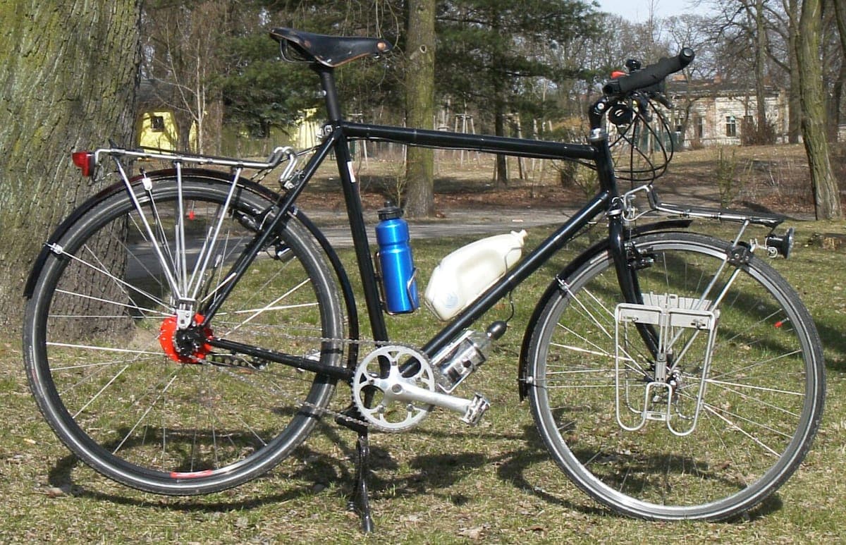 Zwarte stalen reisfiets met rode naafversnelling, voordrager, bagagedrager en blauwe bidon, geparkeerd tegen een boom in een park