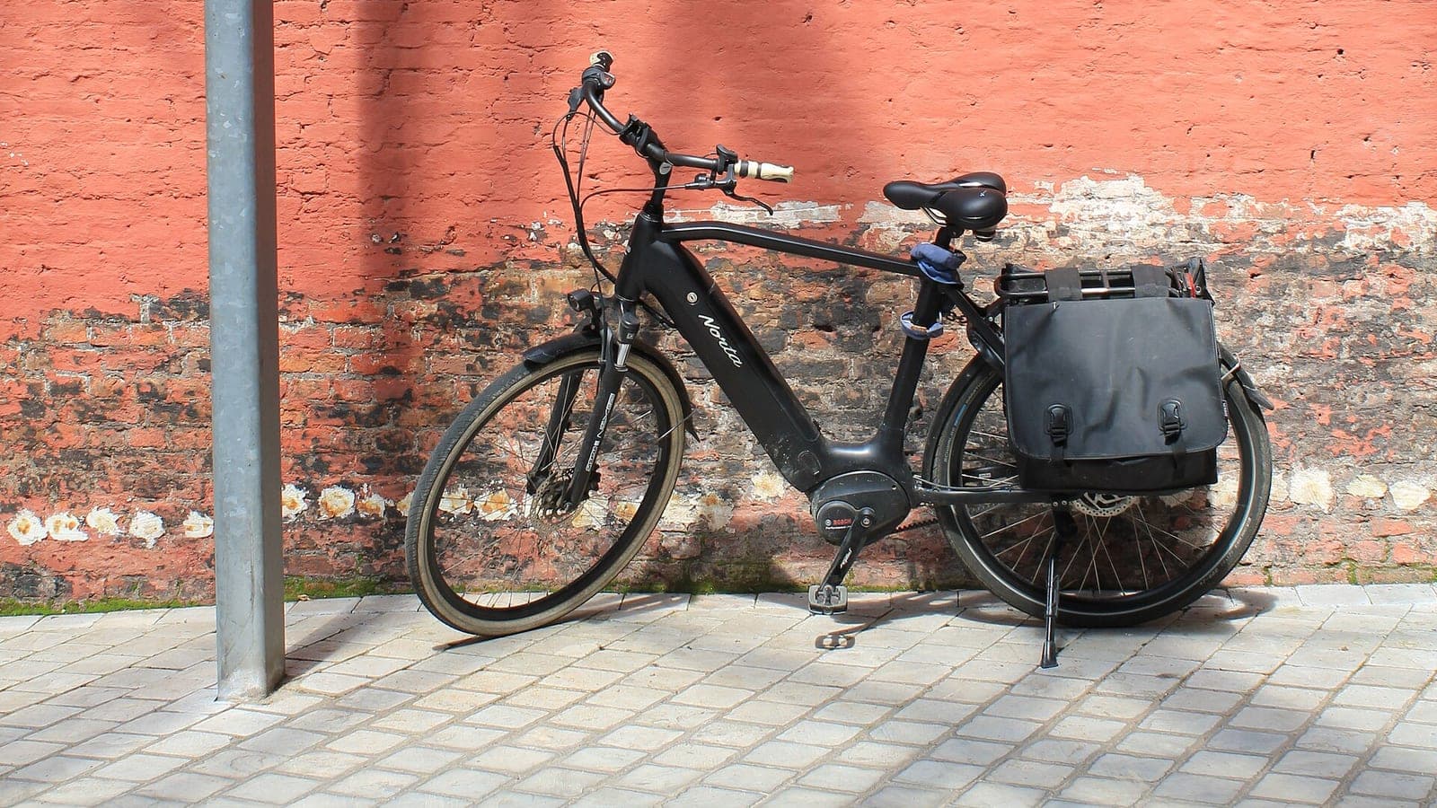 Zwarte elektrische fiets met middenmotor en dubbele fietstassen tegen een oranjerode bakstenen muur